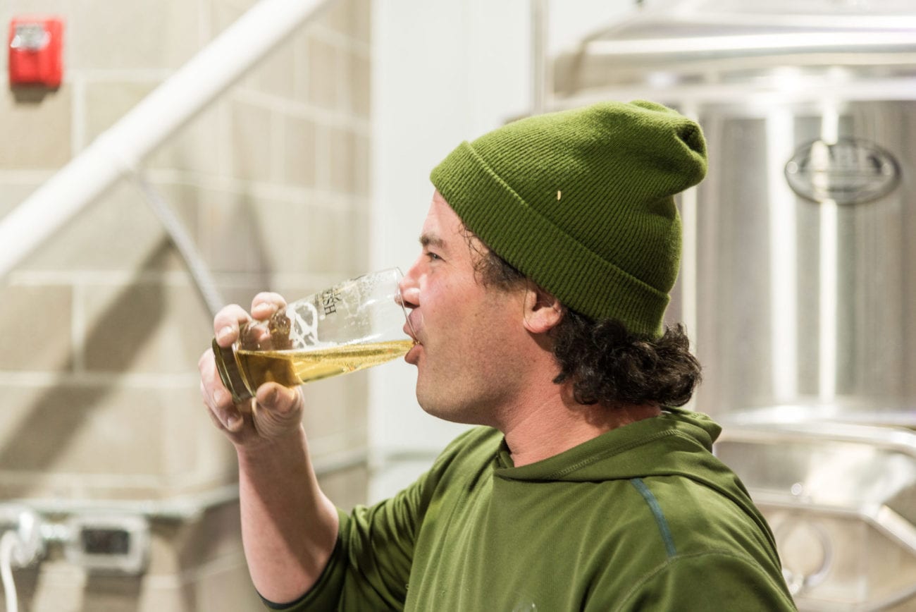 Brewmaster, Nathan Venner, drinking a locally crafted light colored beer with stainless steel brewing equipment in the background.